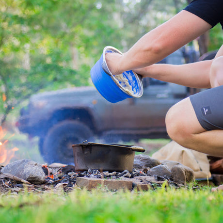 Man scooping ginger beer damper batter from blue bowl above campfire